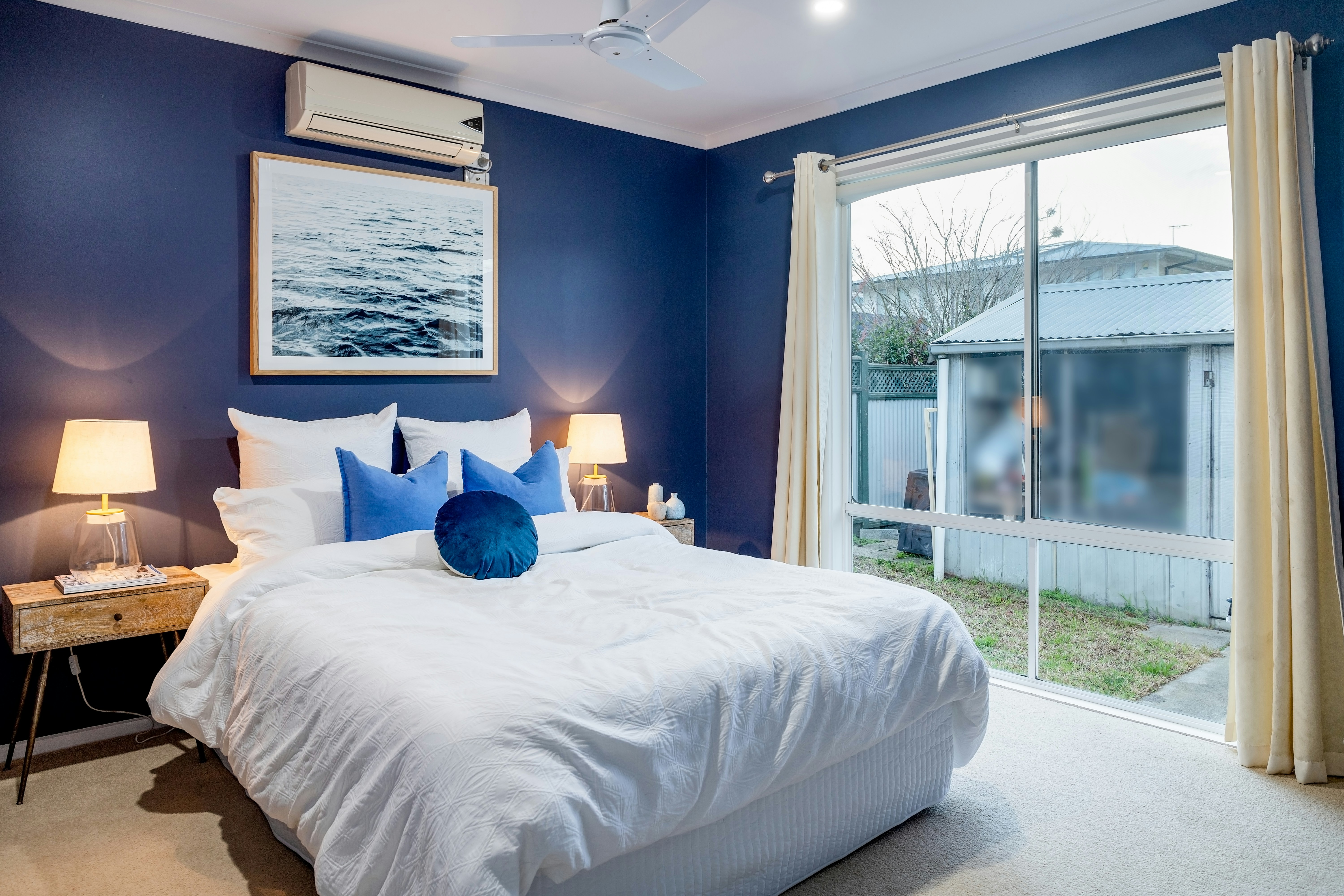 Bright bedroom with navy blue accent wall, white bedding, blue pillows, bedside lamps, and large window with cream curtains.