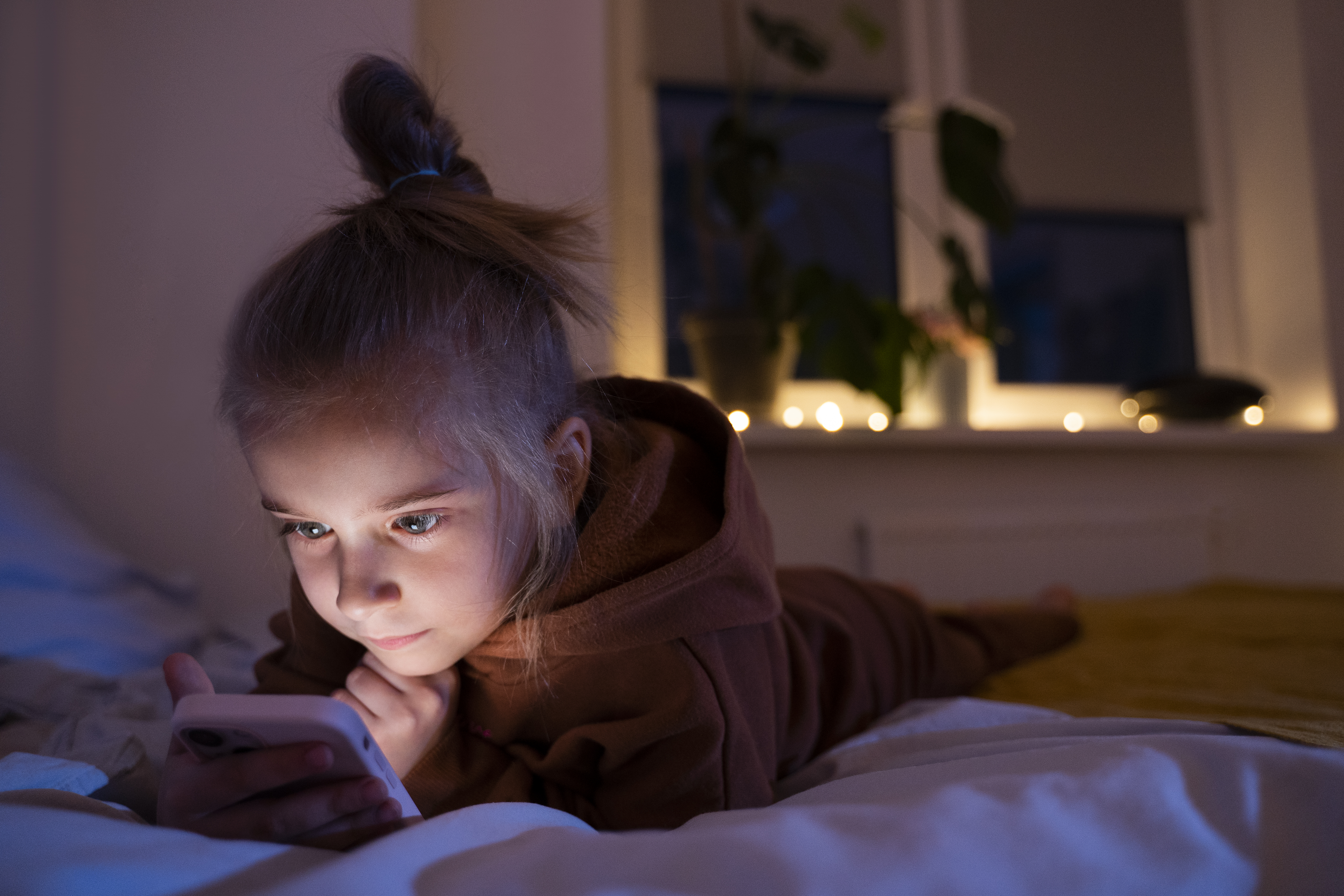 Child lying in bed at night using a smartphone, with screen light illuminating her face.