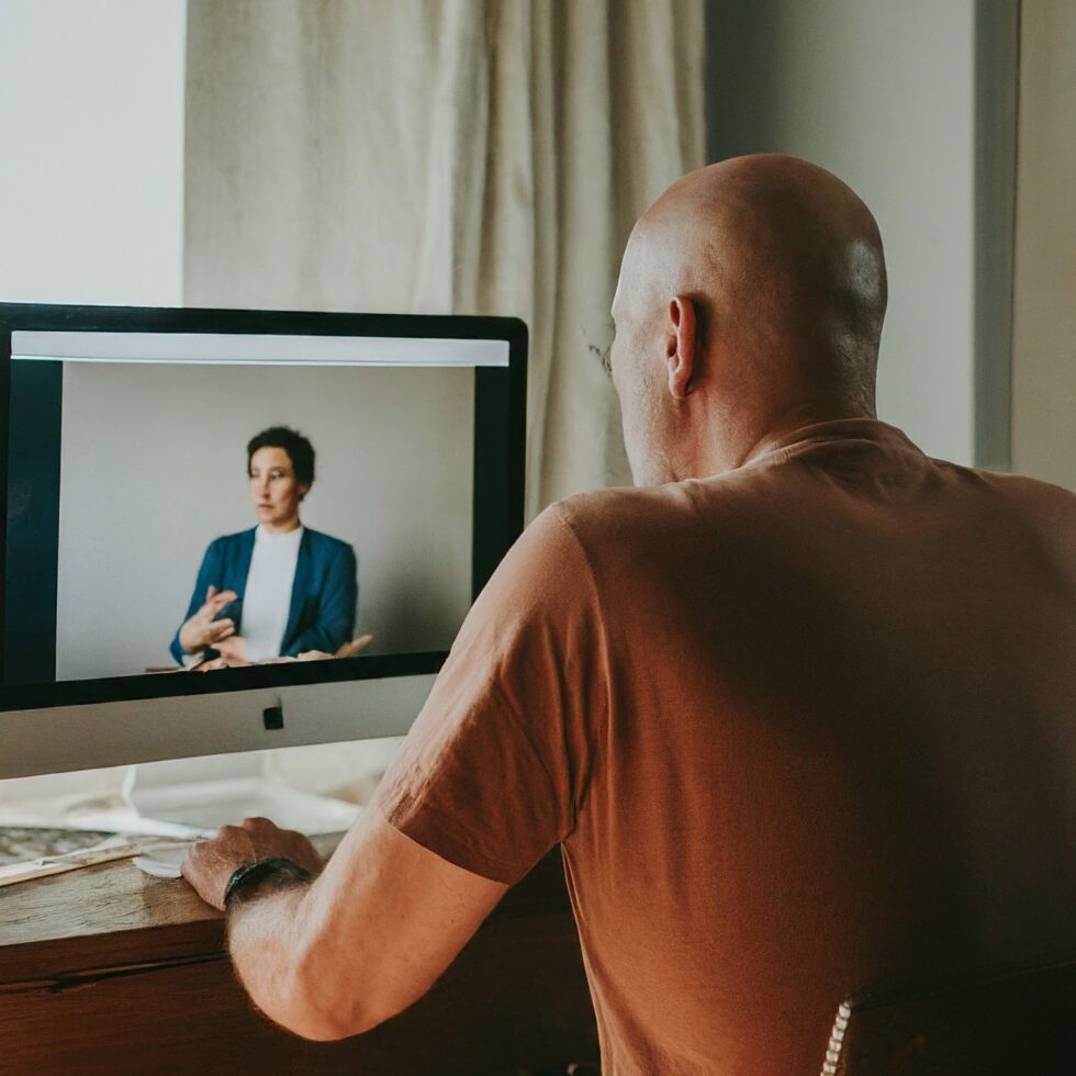 Middle-aged man attending a virtual coaching or therapy session with a specialist on a desktop computer at home.