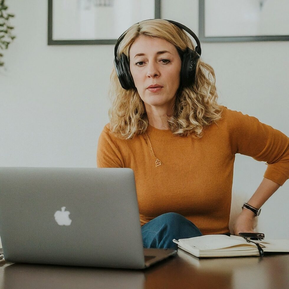 Middle-aged woman wearing headphones during a virtual sleep coaching session on a laptop, with a notebook open beside her.