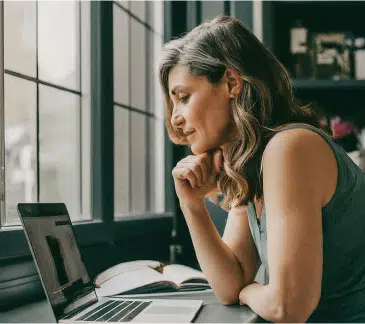 Woman watching an online sleep coaching session on a laptop while sitting by a window with a notebook open.