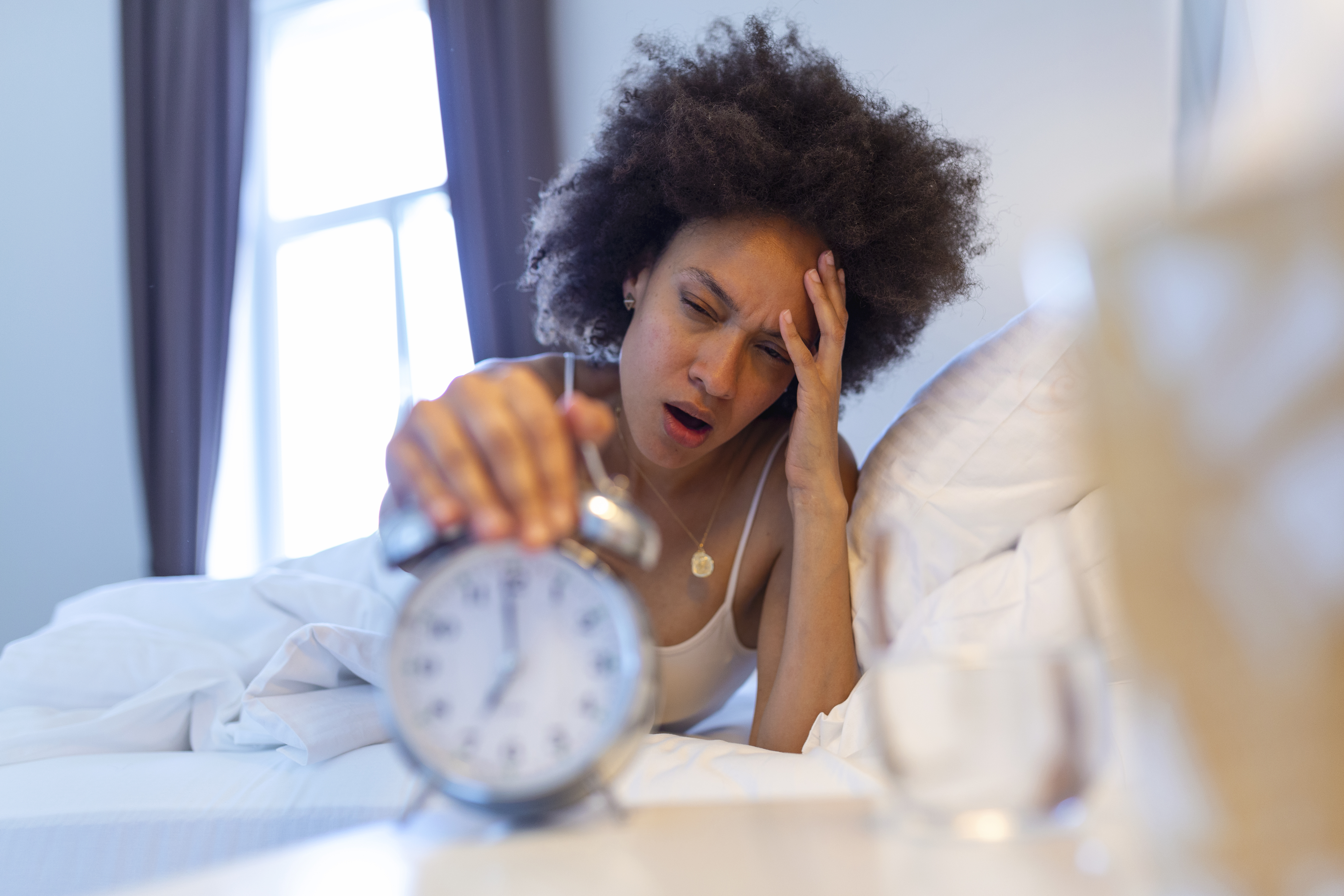 Woman in bed reaching to turn off an alarm clock in the morning, looking tired after waking up early in a bright bedroom
