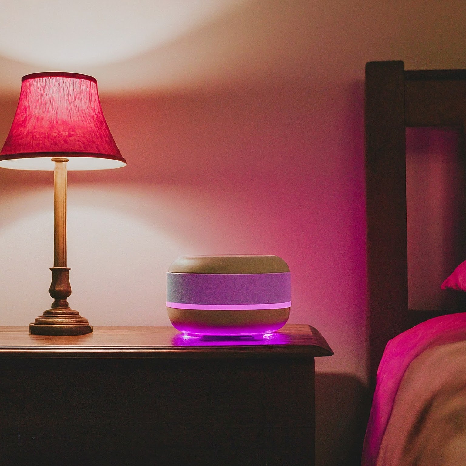 Pink noise machine glowing on a bedside table beside a lamp in a bedroom.