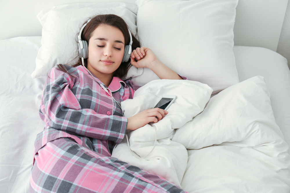 Young woman sleeping in bed while wearing headphones and holding a phone, suggesting relaxing audio before sleep.