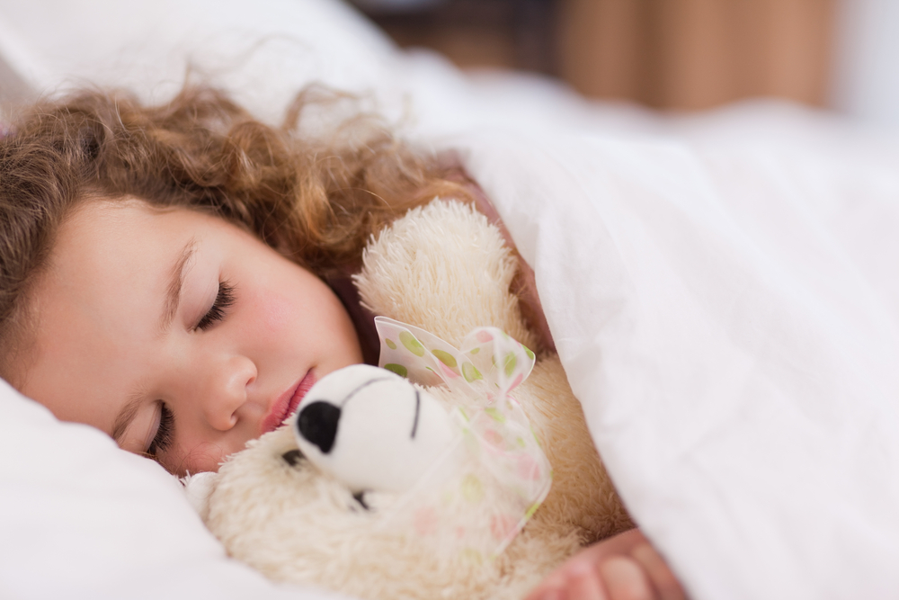 Young child sleeping in bed while cuddling a teddy bear under white blankets.